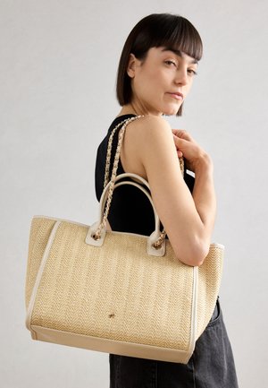 Femme aux cheveux courts et foncés portant un haut noir sans manches tient sur son épaule un grand sac cabas beige tissé avec des anses en chaîne.