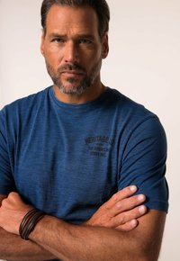 Krátký rukáv modré tričko s kulatým výstřihem, s černě potištěným logem na hrudi. Model má paže zkřížené a nosí kožené náramky.