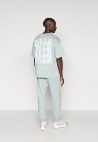 Līdzenzaļš pārmērotais T-krekls ar baltu tekstu uz muguras, saskaņots ar atbilstošām sporta biksēm un gaišas krāsas sporta apaviem.