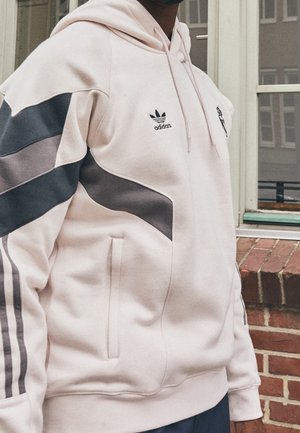 Personne portant un sweat à capuche Adidas rose clair avec des accents gris foncé et des logos, debout près d’un mur en briques et une fenêtre.