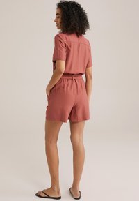 Kortärmad, rostfärgad jumpsuit med bälte i midjan, elastiska sidor och sidofickor. Mjukt, lätt tyg med en avslappnad passform.