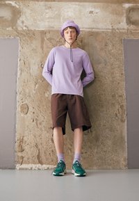 Ljuslila långärmad skjorta, bruna shorts, gröna sneakers, lavendelfärgade strumpor och en lavendelfärgad bucket-hatt med dragsko, framför en texturerad vägg.