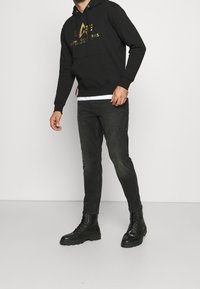 Černá mikina s zlatým logem, zkombinovaná s tmavými džínami a černými šněrovacími botami. Tento outfit vyniká ležérním, městským stylem.