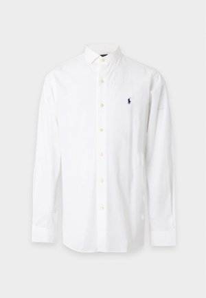 Homme debout portant une chemise blanche à boutons avec un logo, un pantalon foncé et des baskets blanches sur un fond clair uni.