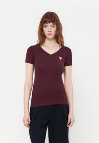 Donna dai capelli rossi che indossa una maglietta attillata a maniche corte di colore bordeaux con una piccola applicazione a forma di triangolo rosso sul lato sinistro del petto, e pantaloni neri.