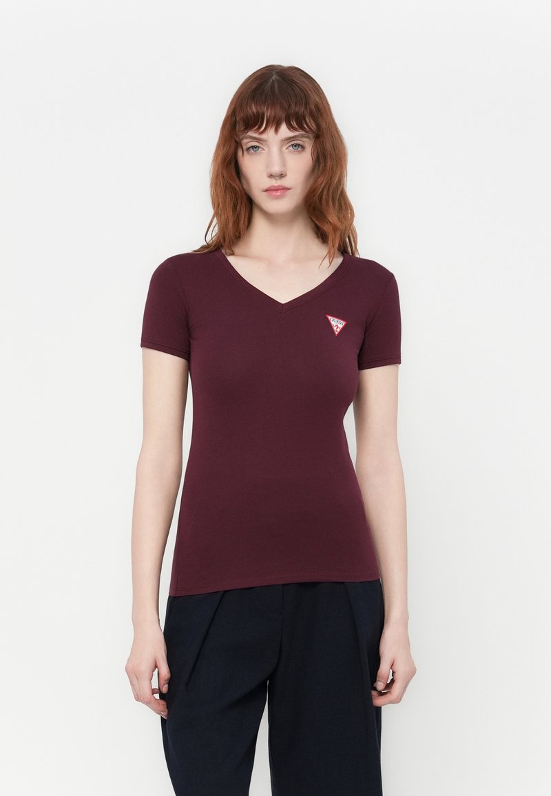 Vrouw met rood haar die een aansluitend bordeauxrood T-shirt met korte mouwen en een V-hals draagt, met een klein rood driehoeklogo op de linkerkant van de borst, en zwarte broeken.