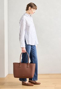 Sac en cuir brun avec deux poignées robustes, de forme rectangulaire, texture lisse et détail minimal en or. Chemise à rayures à boutons et denim.