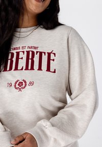 Femme portant un sweat-shirt beige clair avec le texte rouge "LIBERTÉ" et "L'AMOUR EST PARTOUT", ainsi qu'un emblème de rose et l'année 1989.