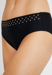 Schwarzer Bikiniunterteil mit goldenen Nietenakzenten entlang des Bundes. Weicher, dehnbarer Stoff in einem niedrig sitzenden Design mit glatter Textur.