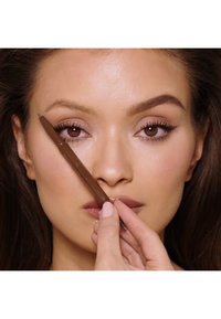 Matita per sopracciglia marrone tenuta vicino al volto di un modello, caratterizzata da un design elegante. Il modello ha un trucco naturale con lunghe ciglia e labbra piene.