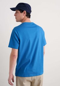 T-shirt din bumbac albastru cu mâneci scurte și guler rotund, văzut din spate. Purtați cu pantaloni bej, asortați cu o șapcă de baseball bleumarin.