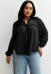 Una donna con lunghi capelli scuri indossa una blusa nera a maniche lunghe con dettagli in pizzo e jeans azzurri. È in piedi contro una parete neutra.