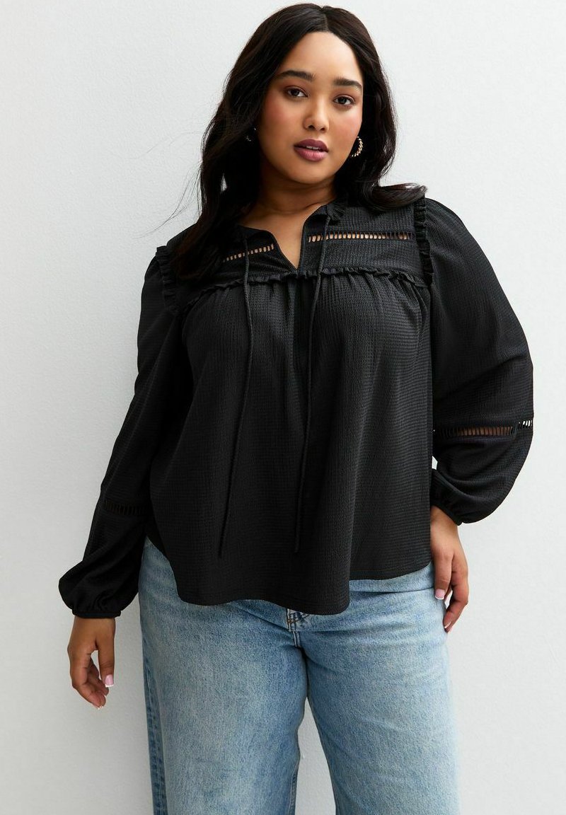 Una donna con lunghi capelli scuri indossa una blusa nera a maniche lunghe con dettagli in pizzo e jeans azzurri. È in piedi contro una parete neutra.