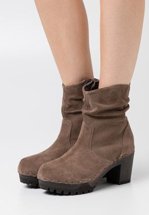 Bottes confortables en daim marron avec un design retroussé, embouts carrés, talons en bois et semelles en caoutchouc comportant des coutures et des accents décoratifs.