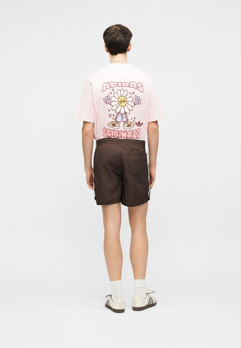 Giovane uomo visto di spalle che indossa una maglietta rosa Adidas Originals con una grafica di un fiore sorridente e pantaloncini marrone scuro.