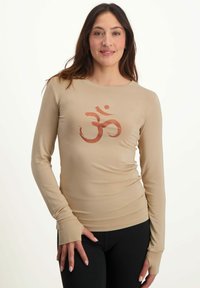 Beige Langarmshirt aus leichtem Stoff mit einem orangefarbenen Om-Symbol-Design in der Mitte. Schlanker Schnitt mit Daumenlöchern an den Bündchen.