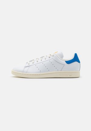 Zapatilla Adidas de corte bajo blanca con suela crema, lengüeta azul en el talón, rayas laterales perforadas y cordones blancos sobre fondo liso.