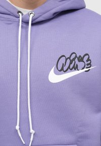 Fialová bavlnená mikina s kapucňou, ktorá má biele logo Nike a čierny text v graffiti štýle na hrudi. Má biele šnúrky a rebrované detaily.