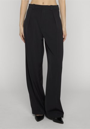 Schwarze weit geschnittene Hose mit hohem Bund, Gürtelschlaufen und vorderen Falten, getragen mit schwarzen spitzen Schuhen, von der Taille bis zu den Füßen modelliert.