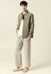 Ljus olivgrön knapp-up jacka med två bröstfickor, vit t-shirt under, beige vida byxor och bruna skor. Slät tygtextur.