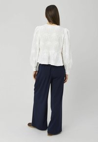 Blouse blanche brodée avec manches bouffantes et ourlet peplum, associée à un pantalon large bleu marine. Texture lisse, motif floral visible.