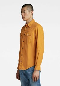 Camicia gialla senape con bottoni neri, dotata di due tasche sul petto, realizzata in un tessuto testurizzato, abbinata a jeans chiari strappati.