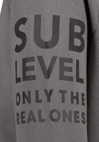 Grauer Hoodie mit auffälligem schwarzen Text, der "SUB LEVEL NUR DIE ECHTEN" liest. Der Stoff hat eine weiche, strukturierte Oberfläche.