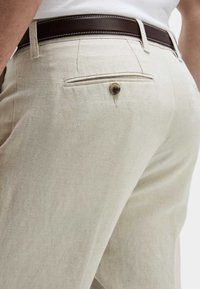 Pantalones de lino color beige claro con un bolsillo trasero con cierre de botón y un cinturón de cuero marrón con costuras blancas. Textura suave.