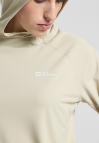 Beige Pullover-Hoodie aus glattem Stoff, mit einem Frontlogo "Jack Wolfskin" und sichtbaren Nähdetails. Kapuze im Hintergrund sichtbar.