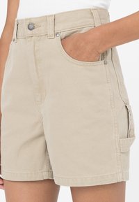 Shorts in cotone beige con vita alta, tasche frontali e una tasca cargo laterale. Il tessuto è strutturato con cuciture visibili e dettagli metallici.