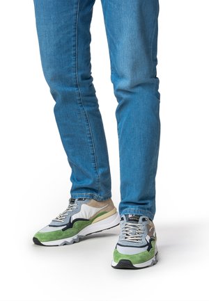 Sportliche Schuhe aus einer Mischung aus grauen, grünen und beigen Materialien; mit strukturierten Wildlederelementen, Schnürverschluss und dicker weißer Sohle. Die Jeans sind blau.