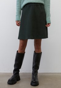 Falda verde oscuro hasta la rodilla hecha de tela suave, con una cinturilla elástica y un diseño simple y recto, combinada con botas negras altas.