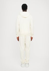 Felpa chiara con cappuccio, dal taglio rilassato e tasca a marsupio, abbinata a pantaloni jogging tapered coordinati. Entrambi presentano polsini e orlo a costine.