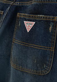 Tkanina dżinsowa w ciemnym niebieskim kolorze z kontrastującym żółtym przeszyciem. Na tylnej kieszeni znajduje się trójkątna czerwono-biała naszywka z logo "GUESS".