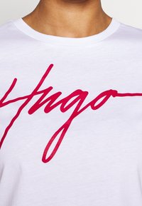Hvid bomulds t-shirt med rund hals, prydet med et stort rødt kursiv "Hugo" logo på forsiden. Blød tekstur, normal pasform.