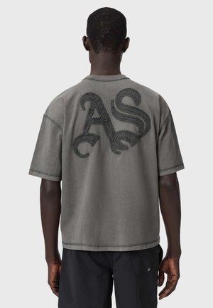 Man draagt een grijs oversized T-shirt met grote, getextureerde "AS"-letters op de achterkant, gecombineerd met een zwarte korte broek.