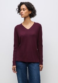 Bordeauxfarbener V-Ausschnitt-Pullover mit langen Ärmeln, geripptem Muster und lockerer Passform. Kombiniert mit hellblauen weiten Jeans.