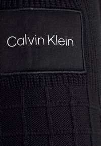 Tessuto a maglia nero con un motivo a griglia, con un'etichetta cucita visibile che mostra "Calvin Klein" in testo bianco.