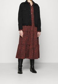 Černá denimová bunda přes červené šaty s květinovým vzorem. Šaty mají víceúrovňový lem, zatímco bunda má klasický límec. Černé kovbojské boty dokončují celý vzhled.