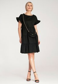 Czarna sukienka z warstwowym dołem i krótkimi, falbaniastymi rękawami, wykonana z lekkiego materiału. Modelka trzyma małą czarną torebkę na ramię. Sandały na obcasie.