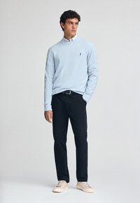 Jasnoniebieski sweter z długim rękawem i kołnierzykiem, noszony na białej koszuli z guzikami. Czarne spodnie i beżowe sneakersy dopełniają stylizację.