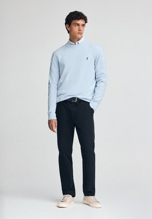 Un pull à manches longues bleu clair avec un col, porté par-dessus une chemise blanche à bouton. Un pantalon noir et des baskets beiges complètent la tenue.