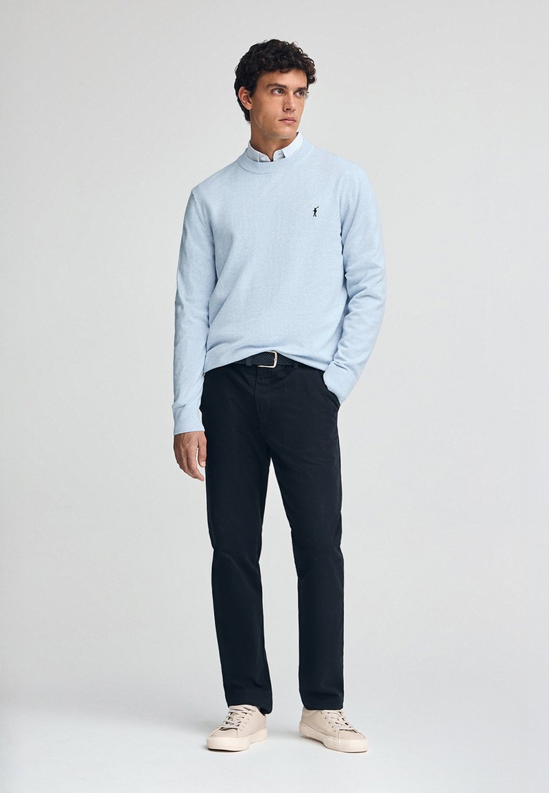 Jasnoniebieski sweter z długim rękawem i kołnierzykiem, noszony na białej koszuli z guzikami. Czarne spodnie i beżowe sneakersy dopełniają stylizację.