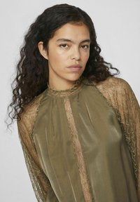 Blouse vert olive avec des manches en dentelle transparente, col montant et plis verticaux. Tissu doux et lisse qui contraste avec des détails en dentelle délicats.