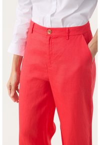 Persoon in helderrode hoge taille broek met knoop en rits, rechterhand in de zak, en een witte lange mouw blouse in de broek gestopt.