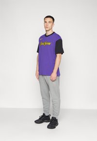 Tricou scurt cu mânecă, de culoare violet și negru, cu grafică "Lakeshow", purtat împreună cu pantaloni scurți gri și adidași negri. Design simplu, croială relaxată.