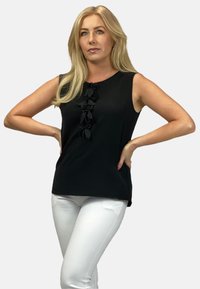 Czarny top bez rękawów z okrągłym dekoltem, zdobiony pionowym rzędem teksturowanych czarnych kwiatów z tkaniny z przodu. W zestawieniu z białymi spodniami.