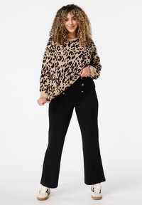Vrouw met krullend haar, gekleed in een sweater met luipaardprint, zwarte geknoopte broek en witte sneakers, staand op een witte achtergrond.