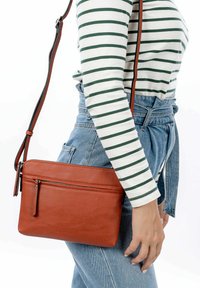 Borsa a tracolla in pelle marrone con superficie testurizzata, dotata di una tasca con zip frontale e di una tracolla regolabile. Posizionata su uno sfondo chiaro.