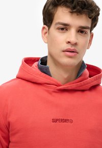 Sudadera roja de algodón con un logo en el frente, que presenta una capucha con cordón y un cuello de mezclilla en capas visible por debajo. Textura suave.